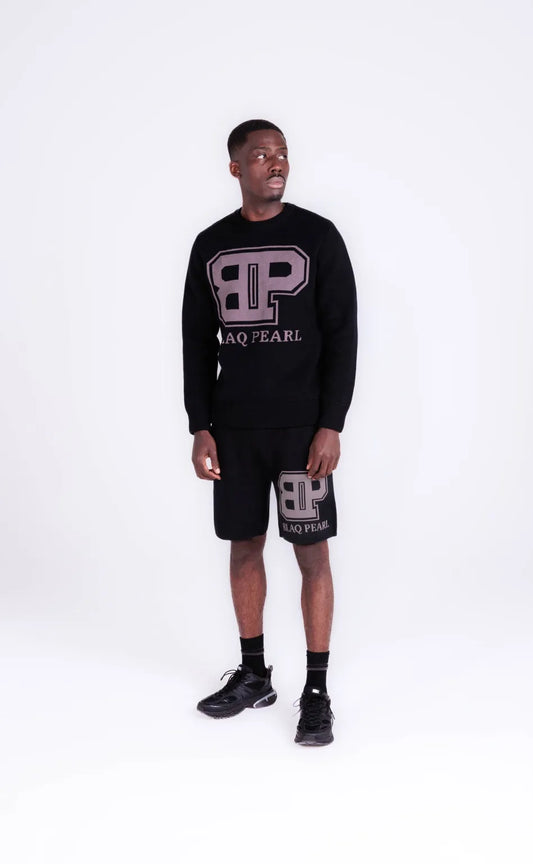 Male model in front view wearing BLAQ PEARL black crewneck jumper with oversized BP logo and matching black shorts – looking to the side, standing against a white background